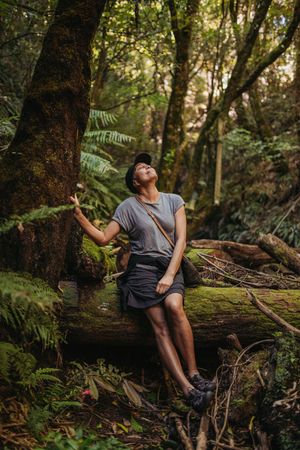 Hiker sitting on mossy log in lush forest, looking up at trees while touching bark, wearing gray tee and black shorts.