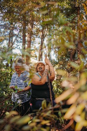 Two women hiking through a lush forest trail, one with tattoos wearing a hat, smiling among green trees.