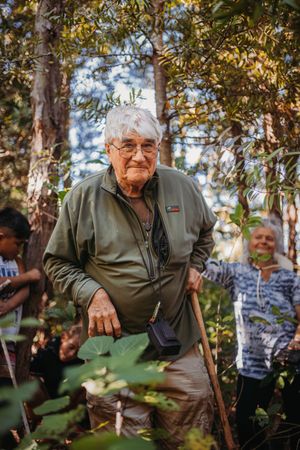 Elderly man with white hair and glasses uses a walking stick while hiking through a lush green forest.