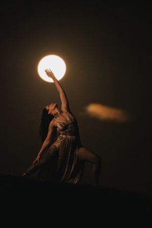 A woman in a flowing dress poses gracefully under a glowing full moon against a dark night sky.