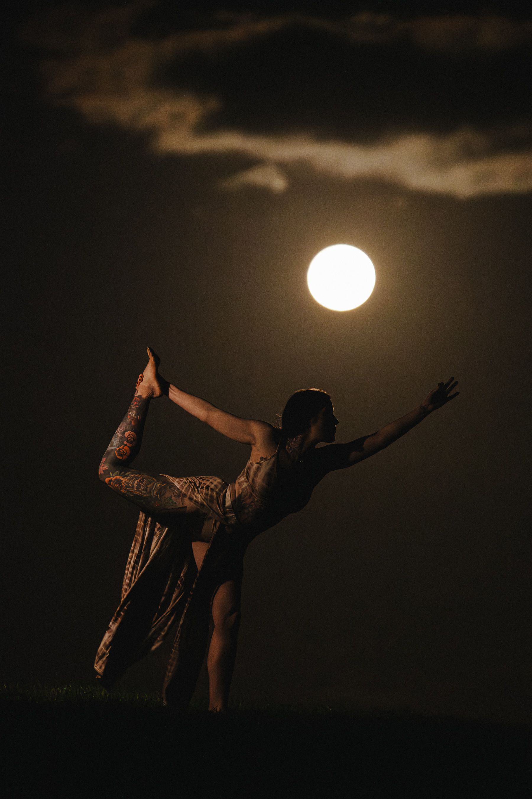 Silhouette of a tattooed yogi performing a dancer pose under a full moon against a dramatic night sky.