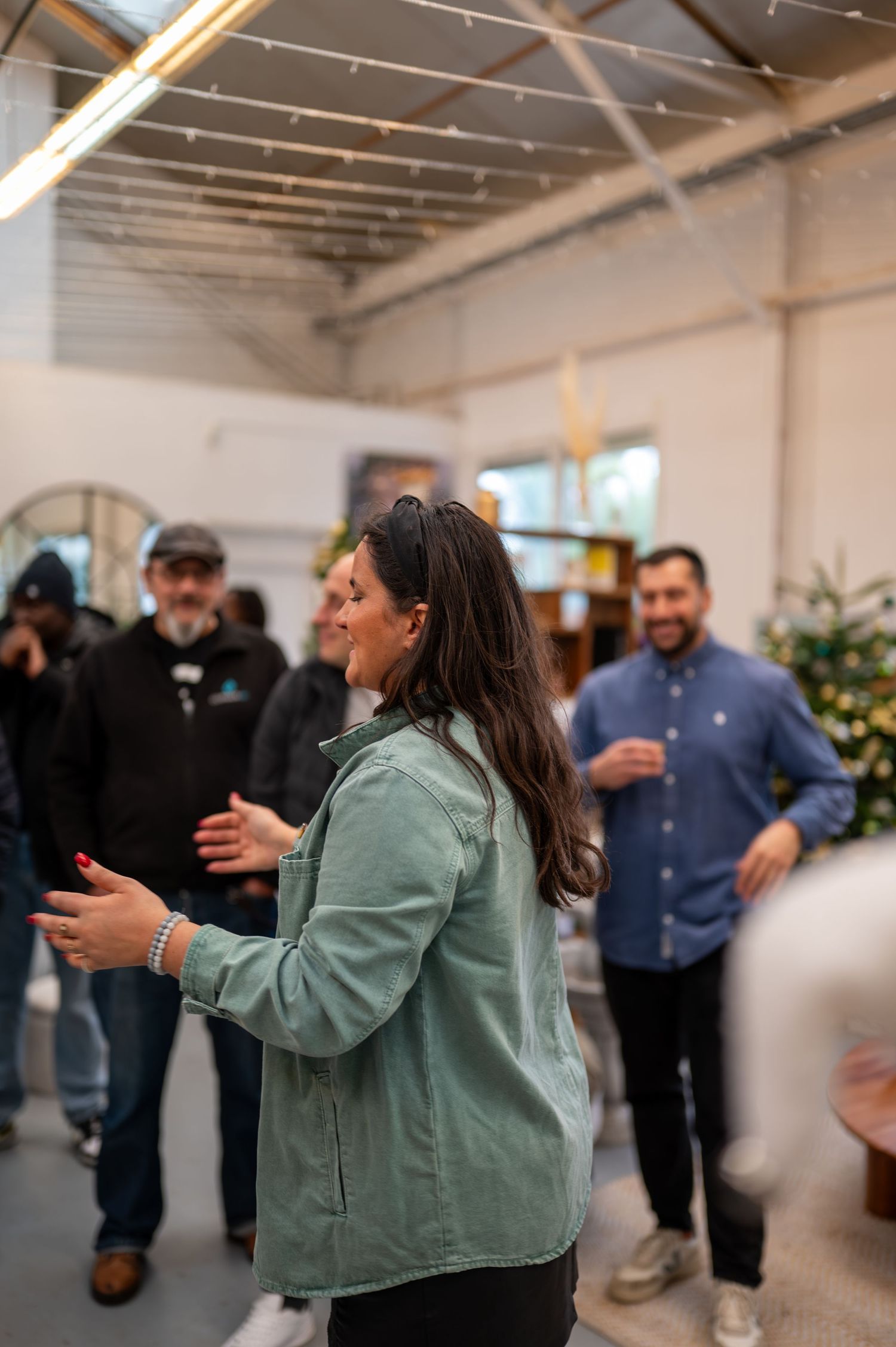 Femme en veste verte applaudissant et souriant lors d'un rassemblement à l'intérieur avec des gens socialisant en arrière-plan.