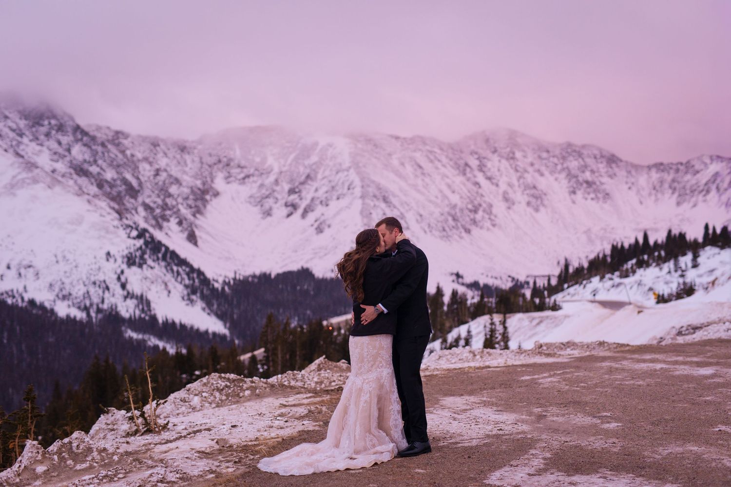 Couple sharing a romantic kiss in wedding attire against a stunning snowy mountain backdrop at sunset.