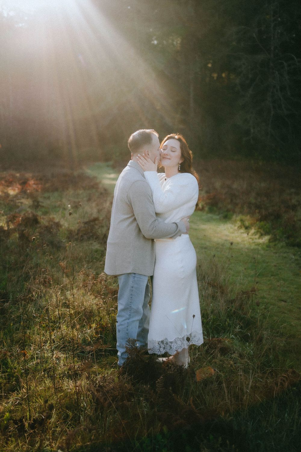 Couple embracing in sunlit forest clearing, bride in white dress and groom in gray suit kissing at golden hour.