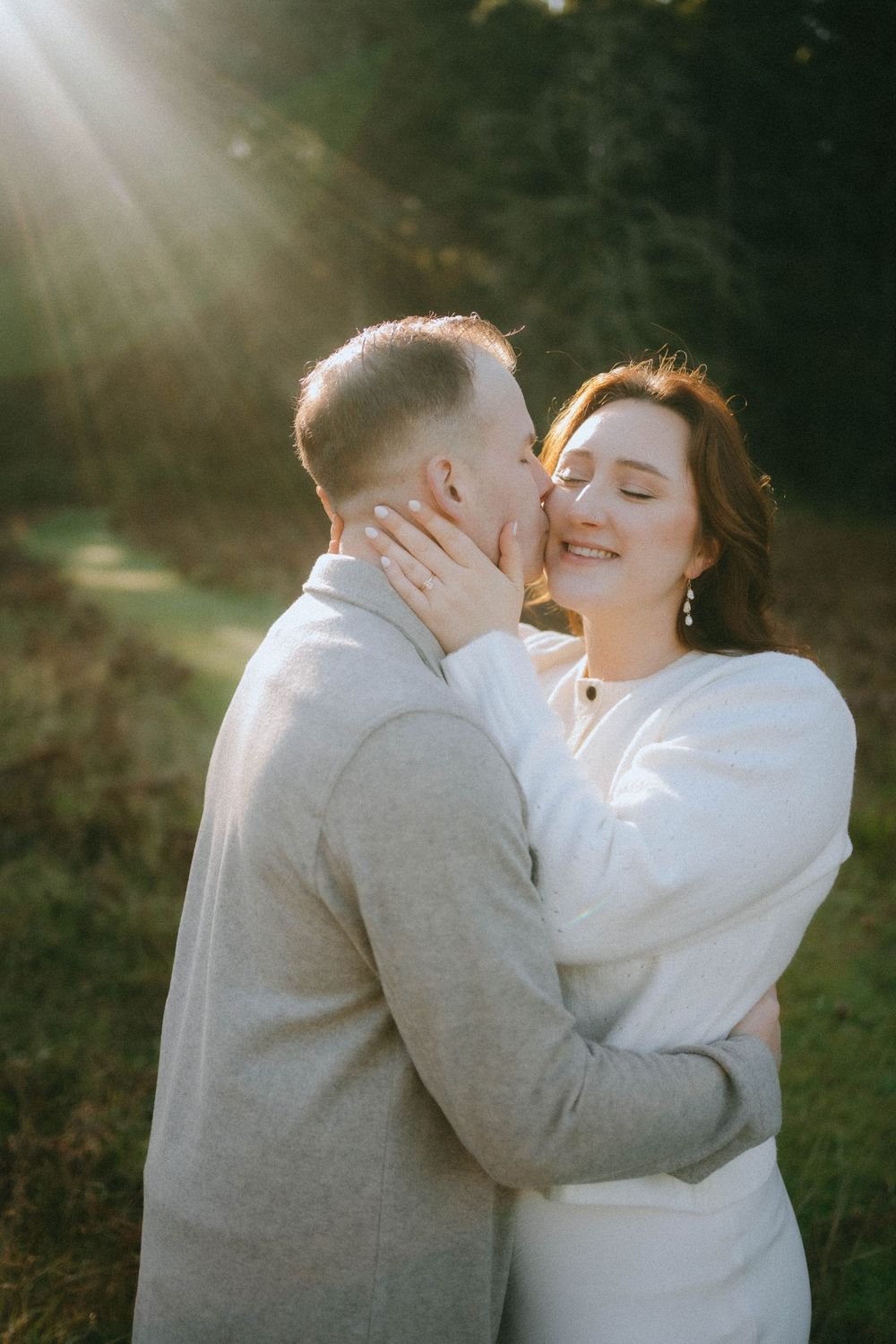 Groom kissing smiling bride on cheek in sunlit woodland, bride in white dress with eyes closed in joy.