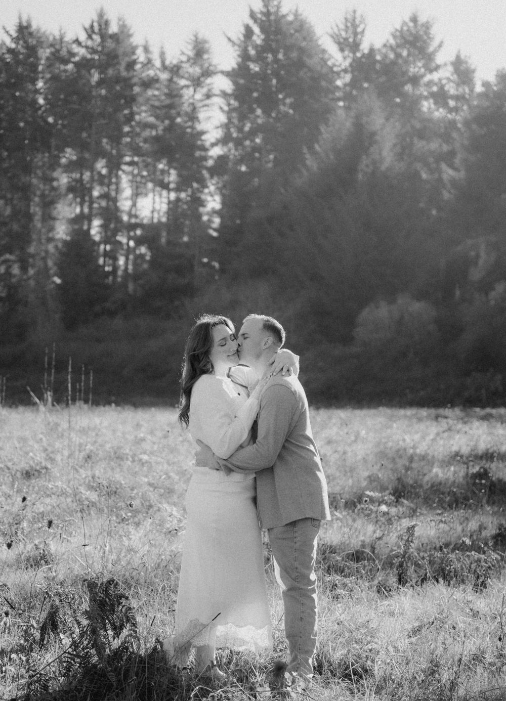 Black and white photo of a couple embracing and kissing in a meadow with tall evergreen trees in the background.