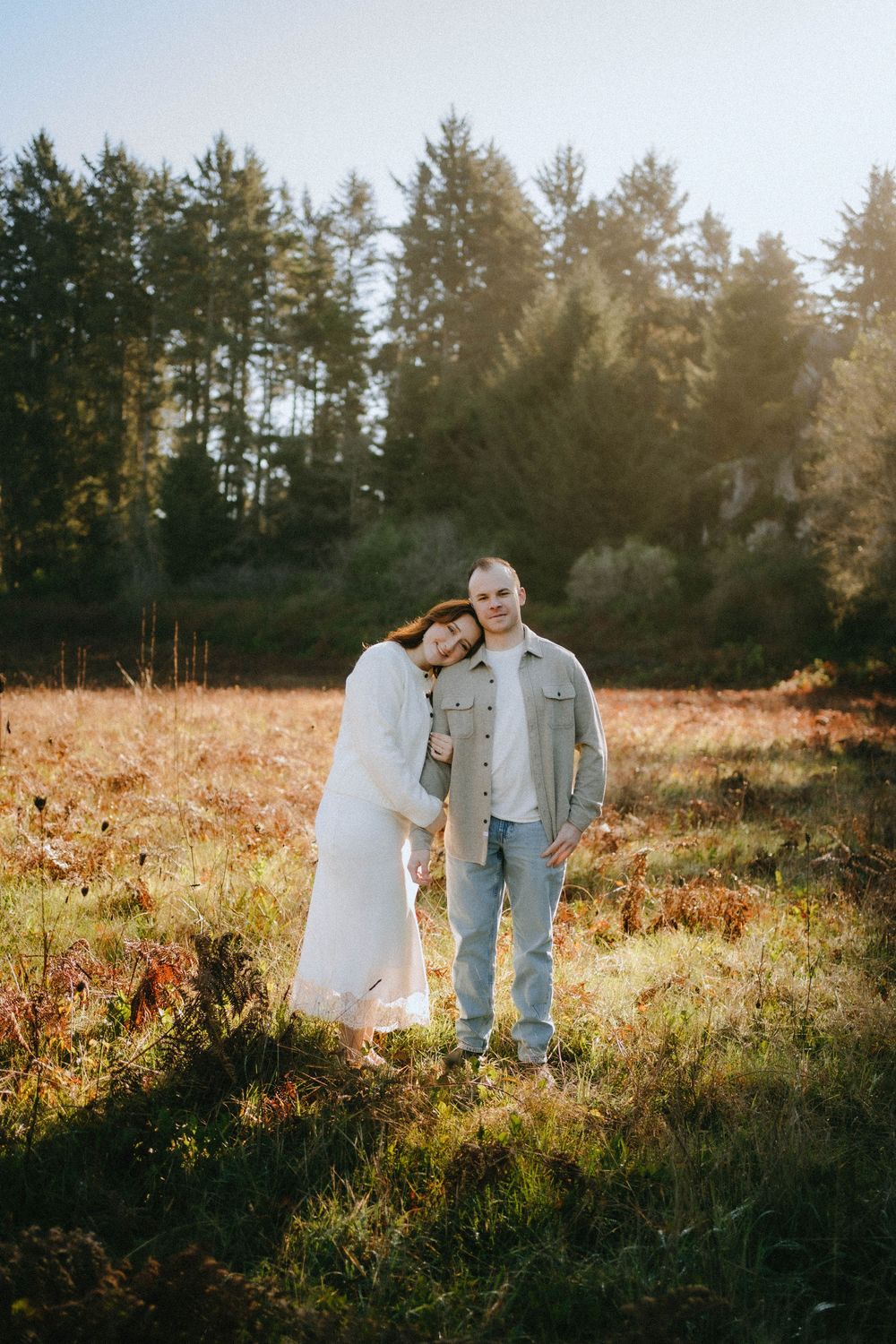 Couple embracing in a sunlit autumn meadow with tall evergreen trees in the background.