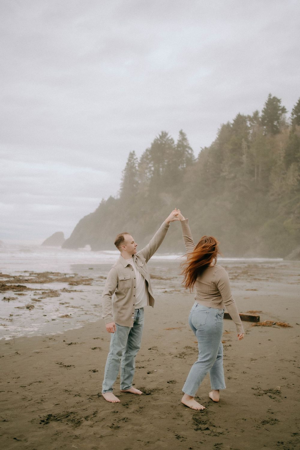 Couple dancing on a misty Pacific Northwest beach, man spinning woman with trees and rocky cliffs in background.