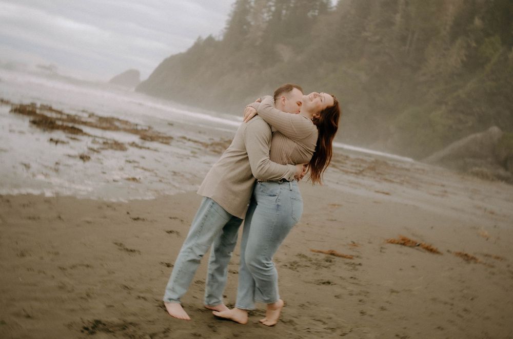 A couple shares a romantic kiss on a misty beach, embracing barefoot on sandy shores with foggy coastal cliffs behind them.
