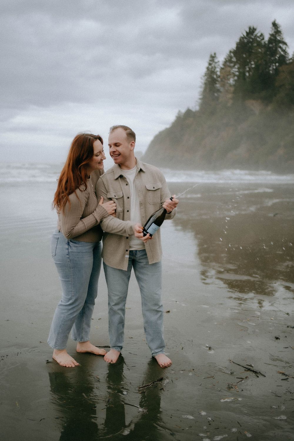 Couple standing barefoot on a misty beach, sharing a bottle of wine, wearing matching beige shirts and jeans.