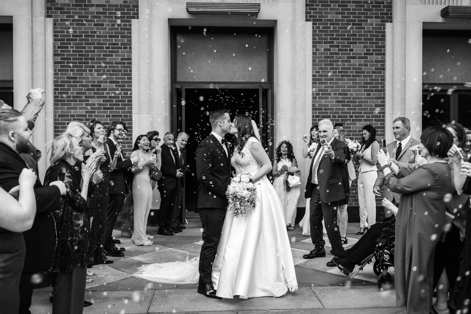 Black and white photo of newlyweds kissing as guests celebrate with confetti outside a brick building.