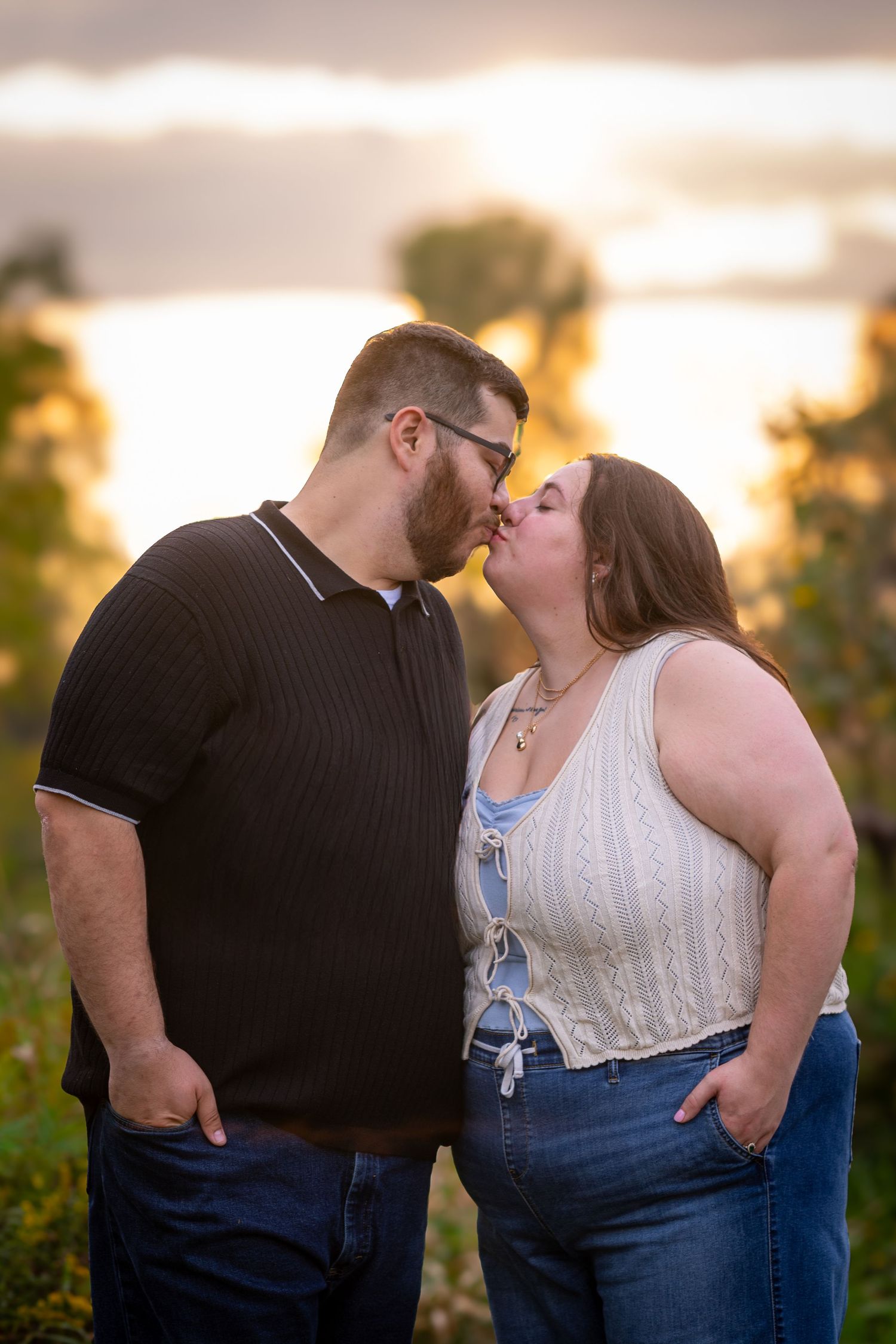 A couple shares a romantic kiss outdoors at golden hour with a glowing sunset sky behind them.
