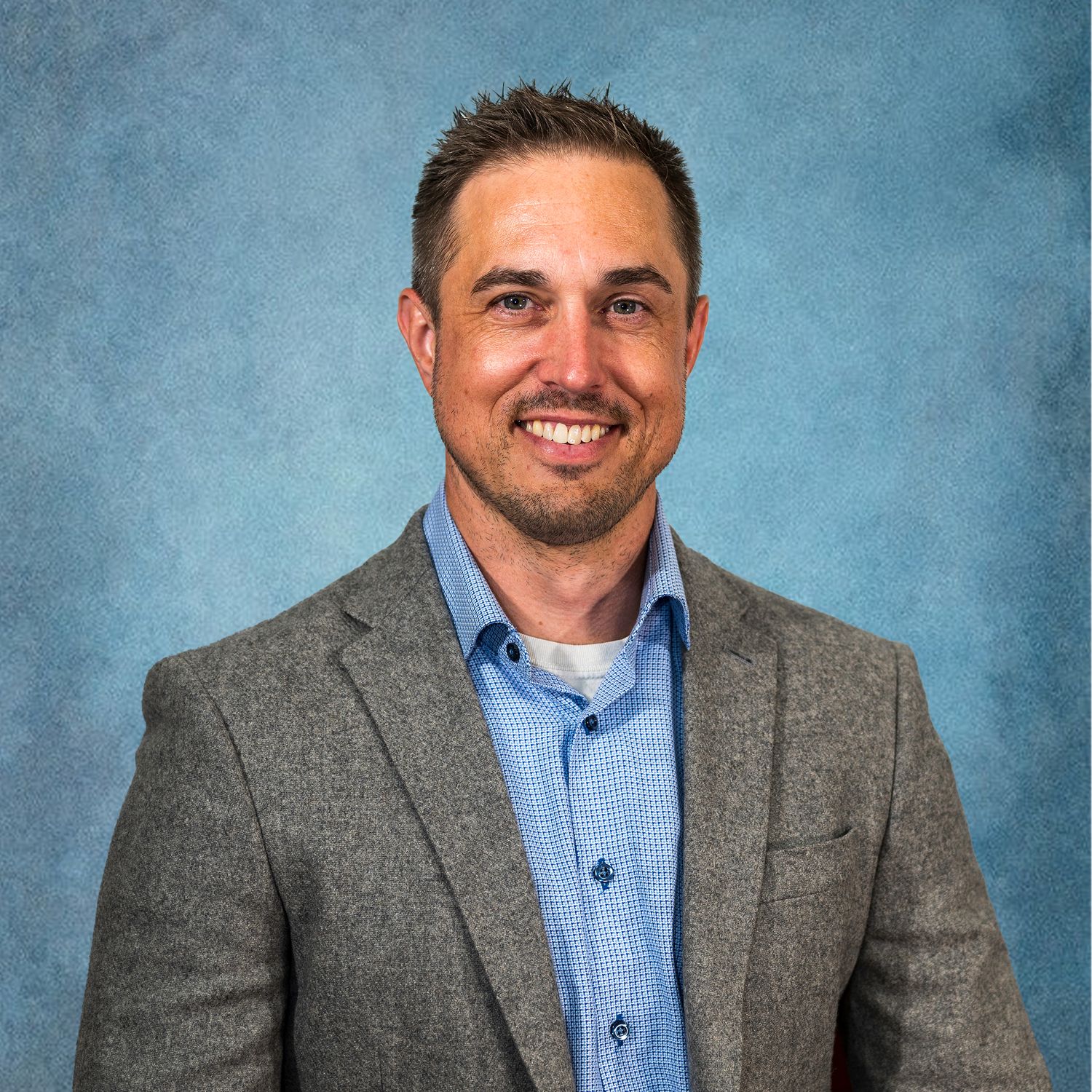 Professional man in gray blazer and blue shirt smiling confidently against a blue studio background.