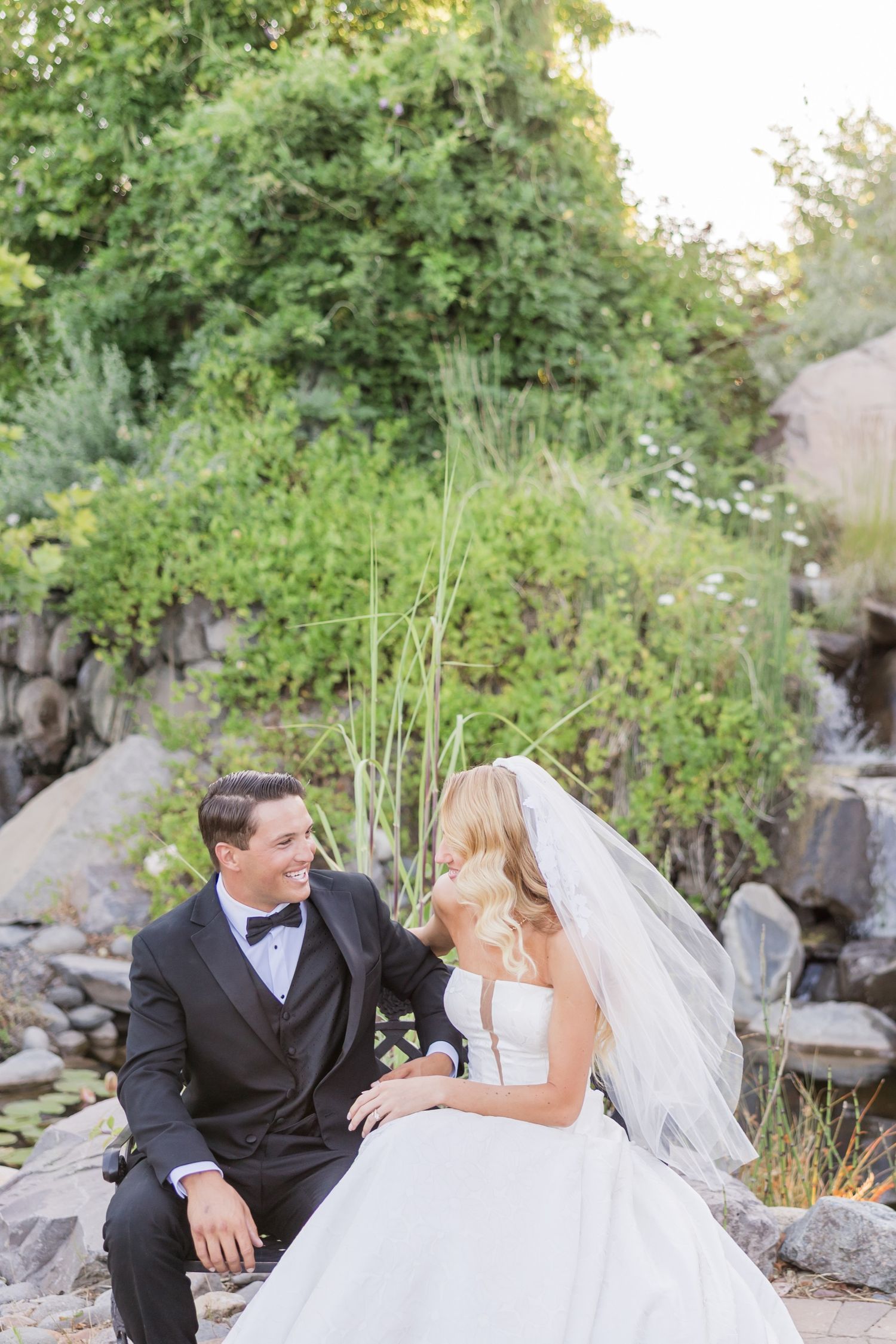 Bride and groom sitting together on rocks near a garden pond, gazing at each other lovingly on their wedding day.