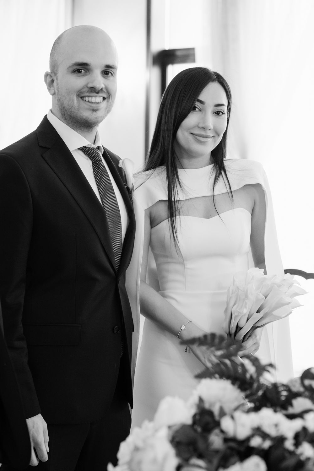 Black and white wedding photo of smiling bride holding white calla lilies bouquet standing beside groom in dark suit.