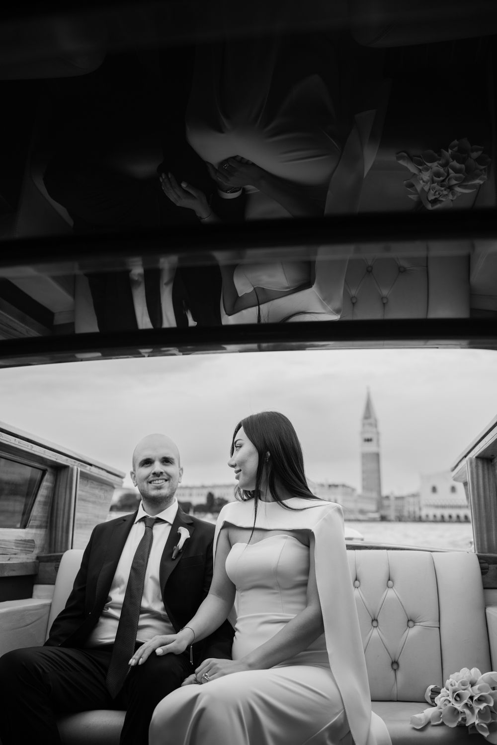 Black and white photo of a bride and groom sitting together on a boat in Venice, Italy, with St. Mark's Campanile in background.