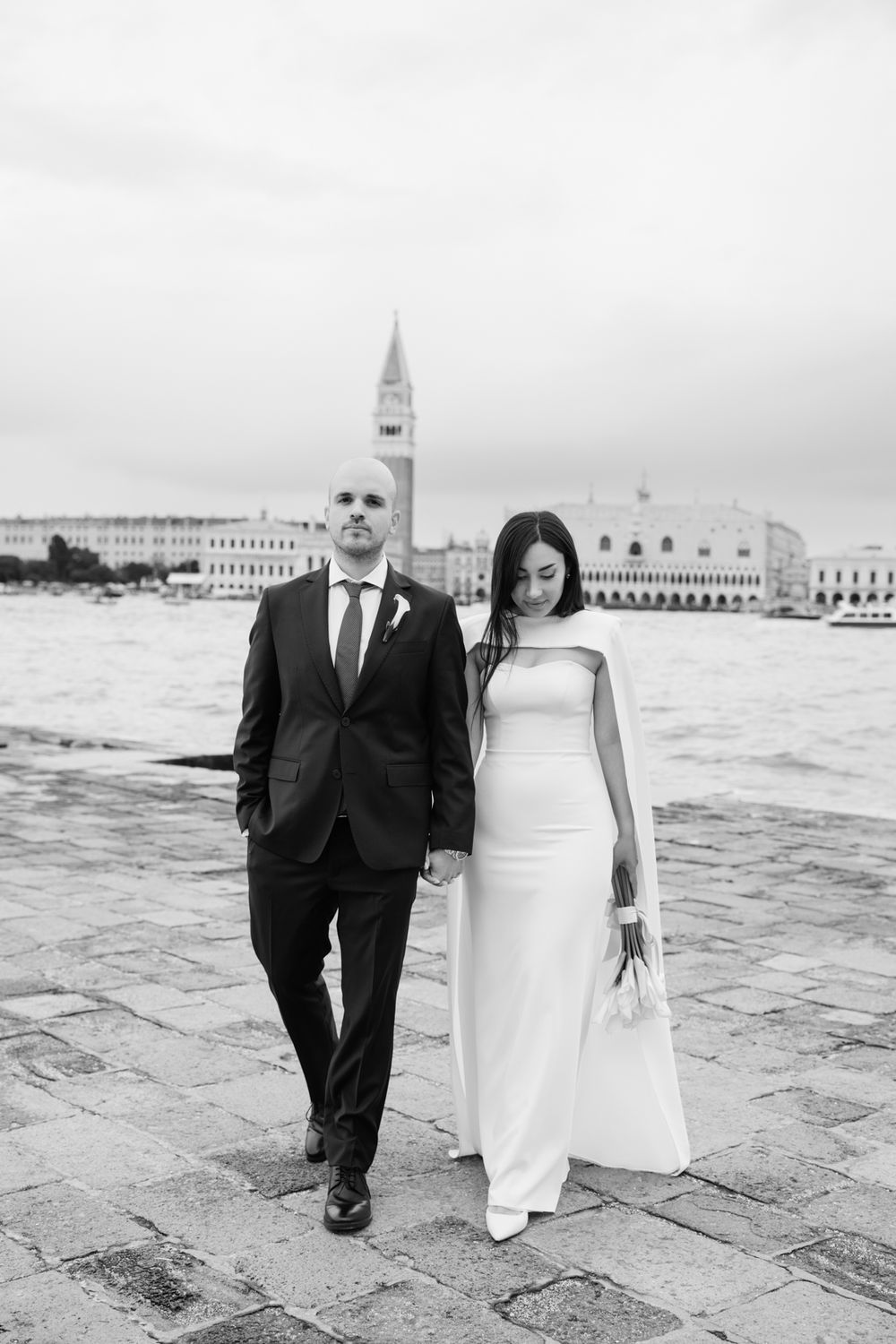 Black and white photo of a bride and groom holding hands in Venice, Italy, with St. Mark's Campanile in the background.