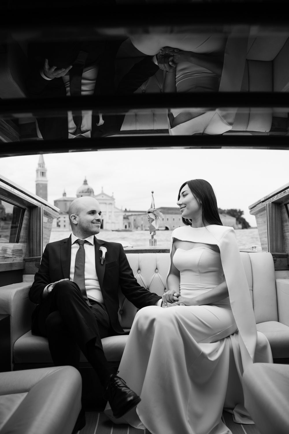 Black and white photo of elegant bride and groom holding hands in a luxury boat with Venice skyline backdrop.