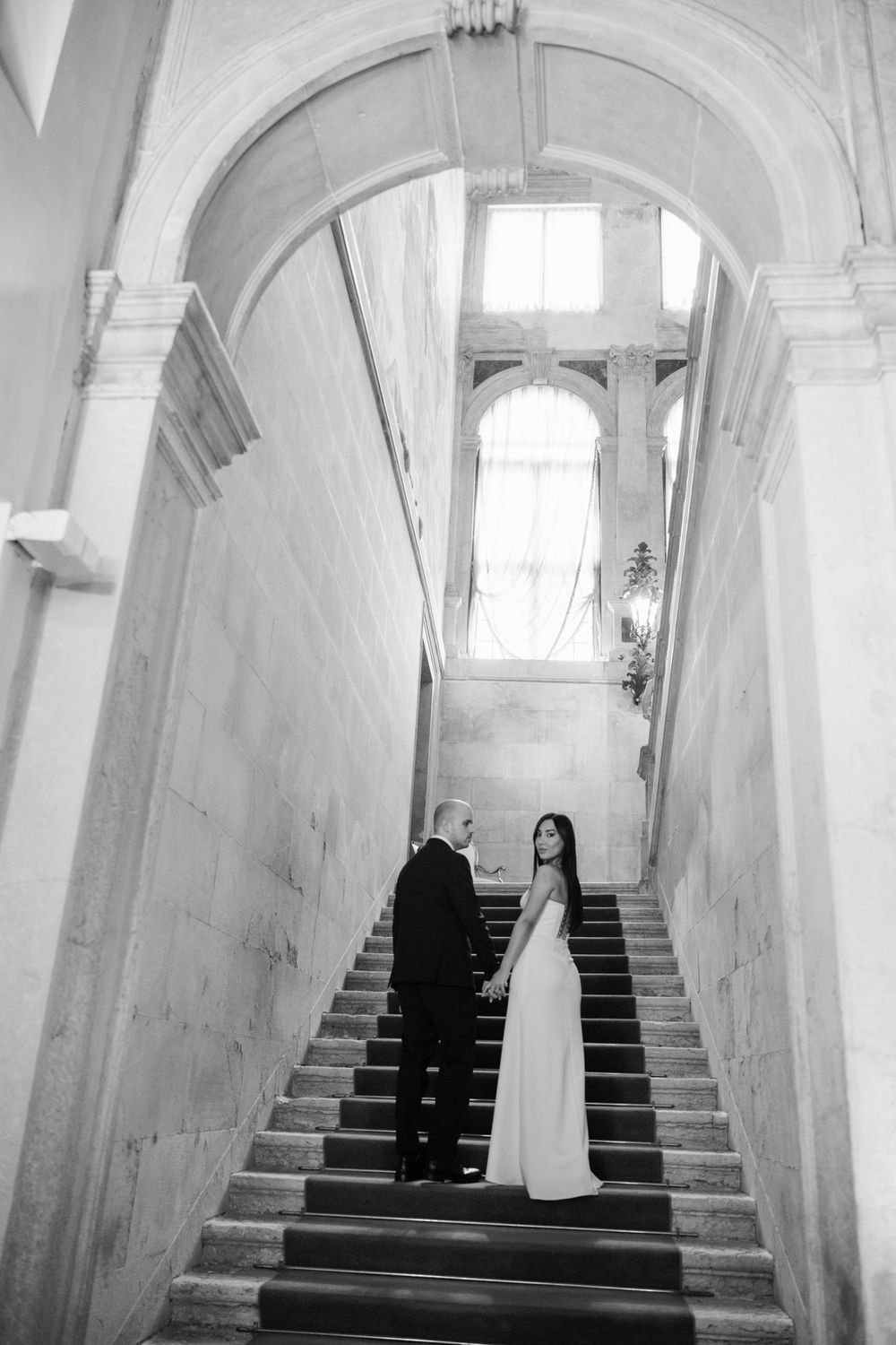 Black and white photo of a bride and groom holding hands on grand marble staircase in elegant venue.