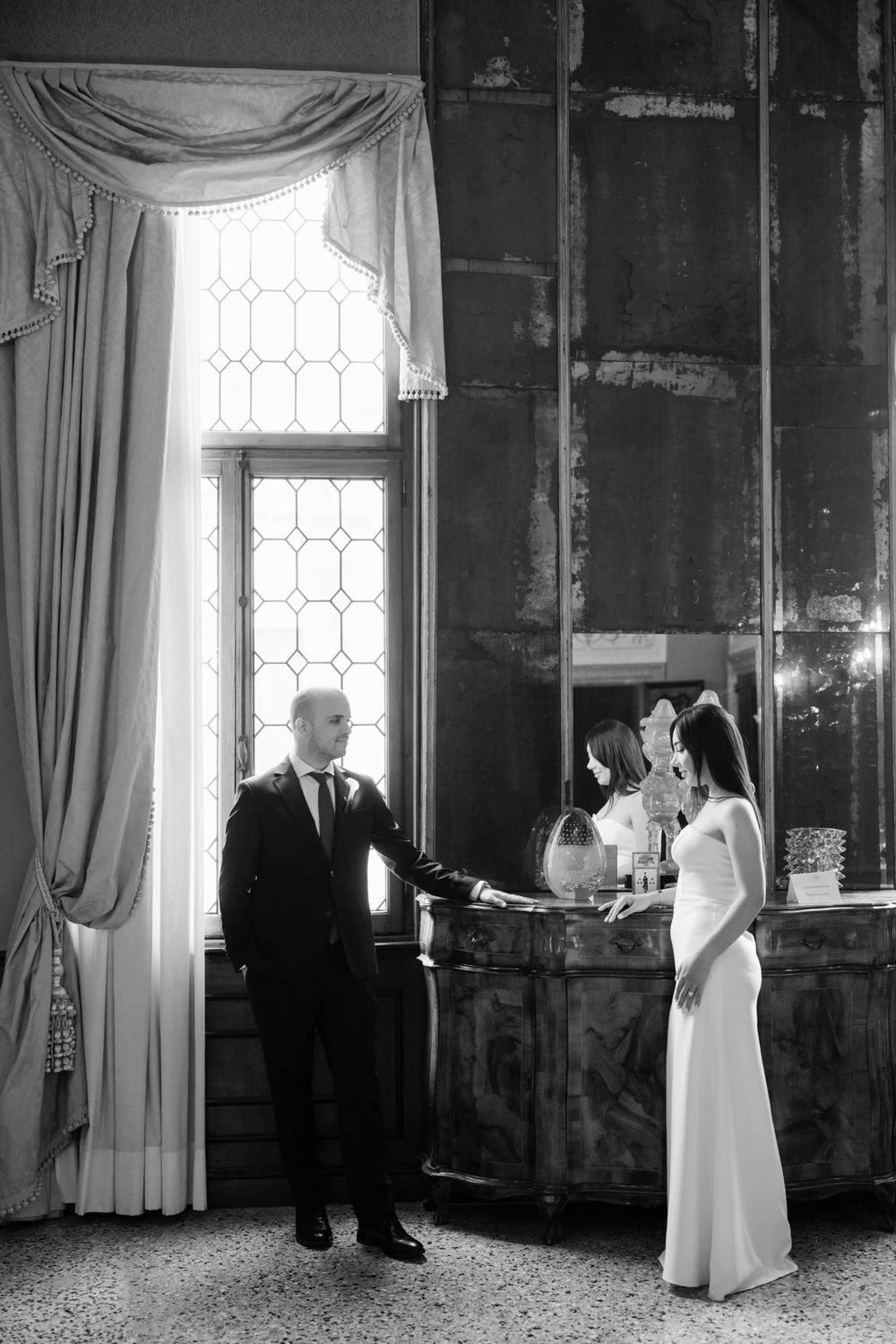 Elegant bride and groom holding hands in a grand black and white wedding portrait by a tall window.