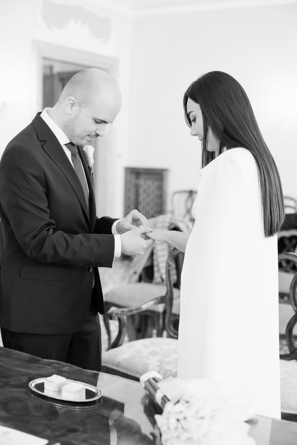 Black and white photo of groom placing wedding ring on bride's finger during elegant indoor ceremony.