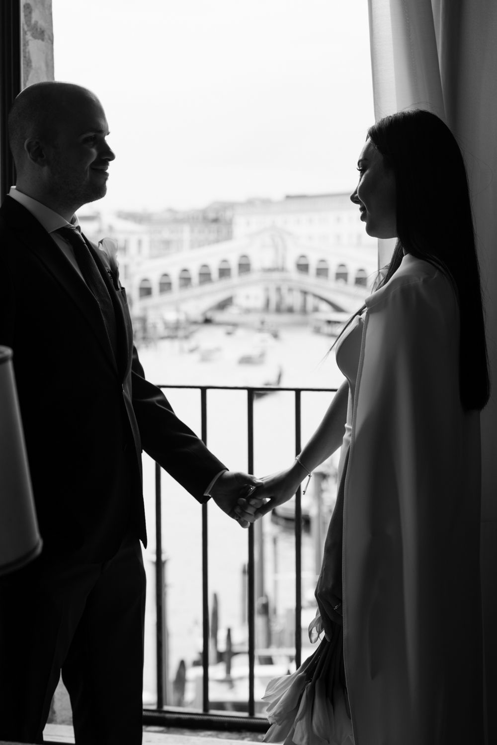 Black and white photo of a couple holding hands by a balcony overlooking the Rialto Bridge in Venice.