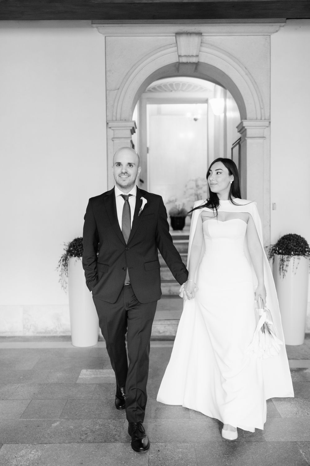 Black and white photo of a bride and groom holding hands walking out of an elegant arched doorway on their wedding day.