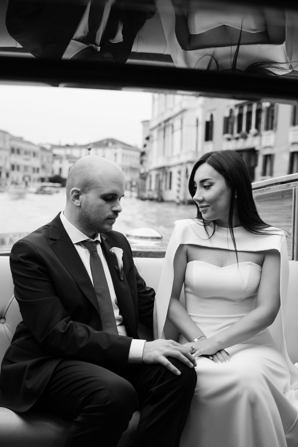 Black and white photo of a bride and groom holding hands on a gondola in Venice, Italy.
