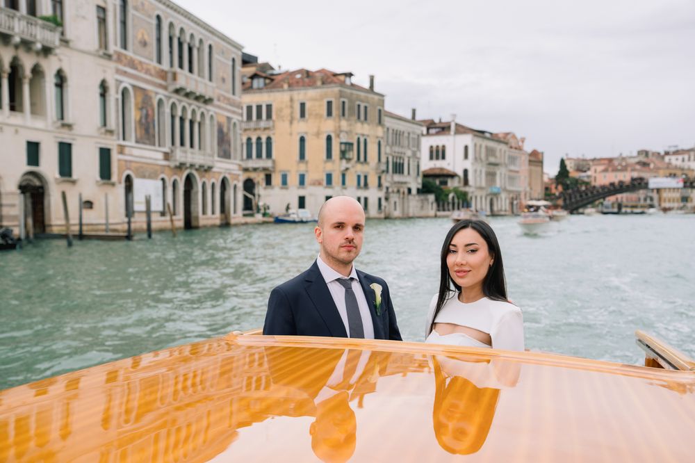 Couple in wedding attire on a wooden boat along Venice Grand Canal with historic buildings in background.