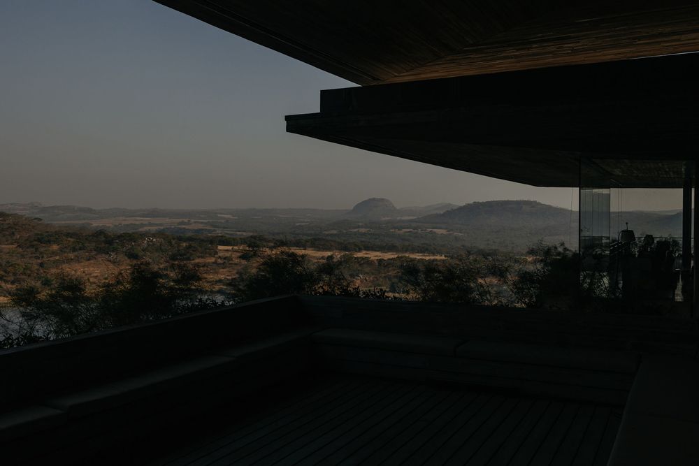 Panoramic view from a shaded deck overlooking a vast African savanna landscape with distant mountains at dusk.