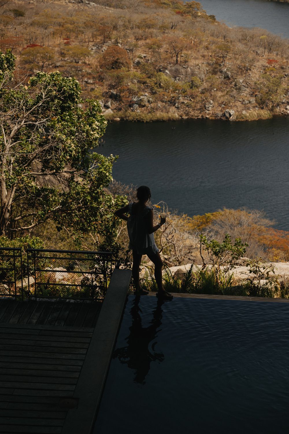 Silhouette of a person standing at an infinity pool edge overlooking a scenic river and dry landscape.