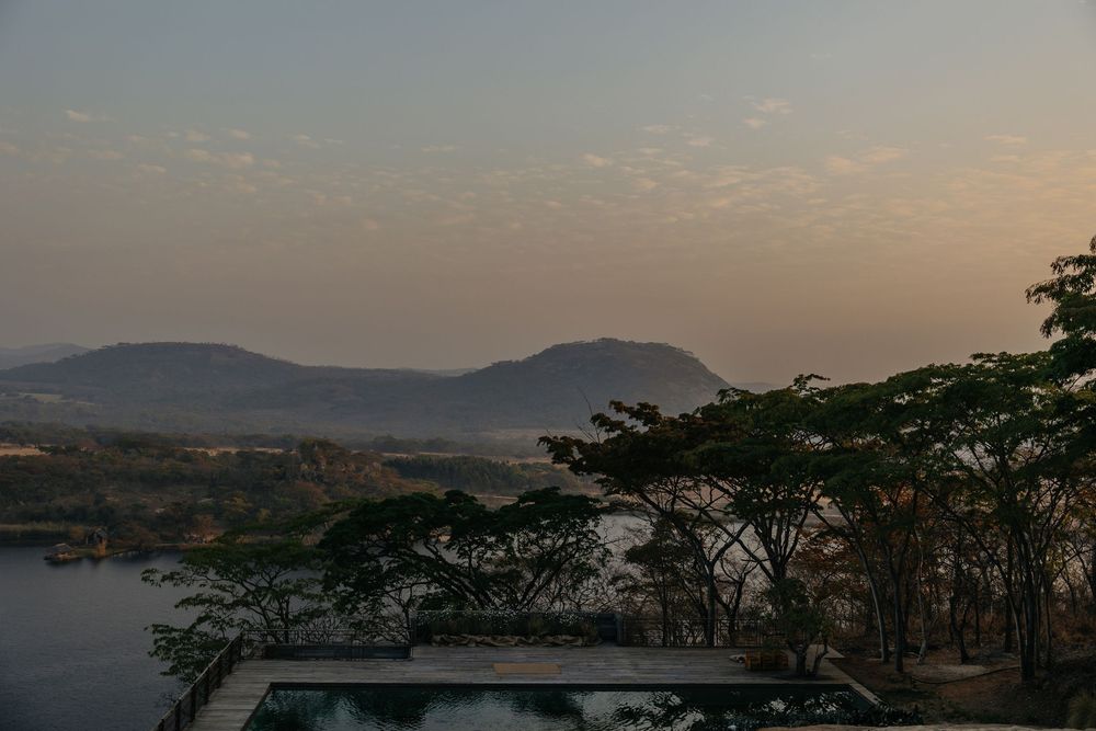 Scenic sunset view over a tranquil lake with rolling hills, acacia trees, and a hazy sky in an African landscape.