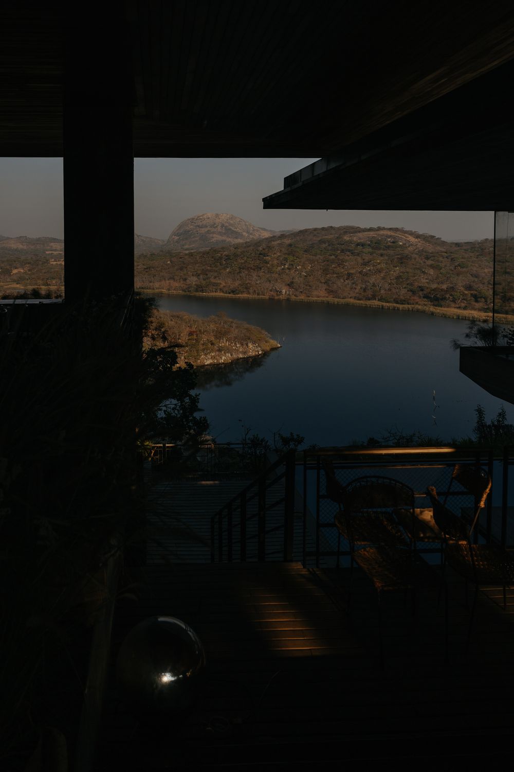 Twilight view of a serene lake surrounded by hills, seen from a dark covered deck with steps leading down.