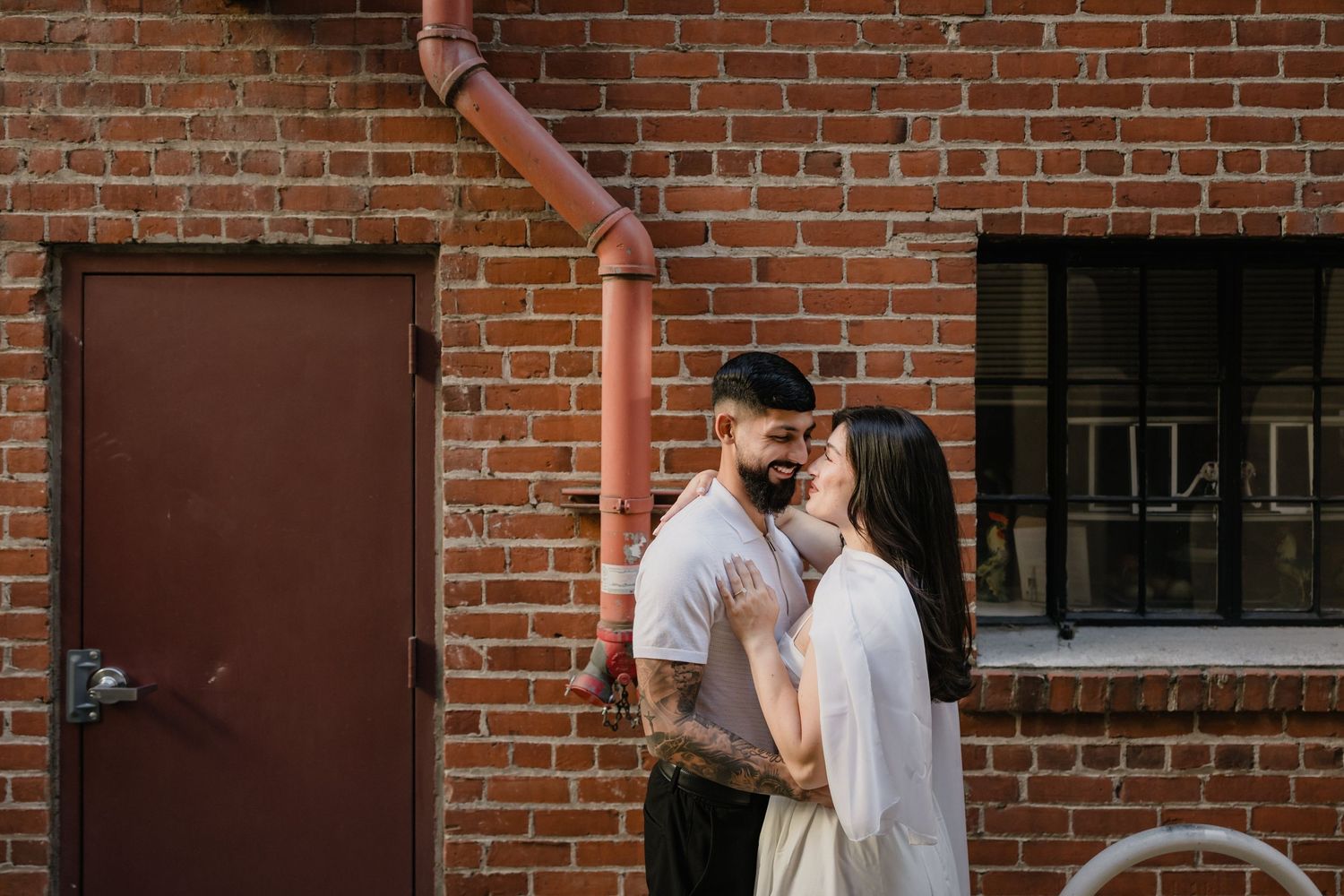 A tattooed couple shares a romantic moment in front of a rustic brick wall with a red pipe and vintage door.