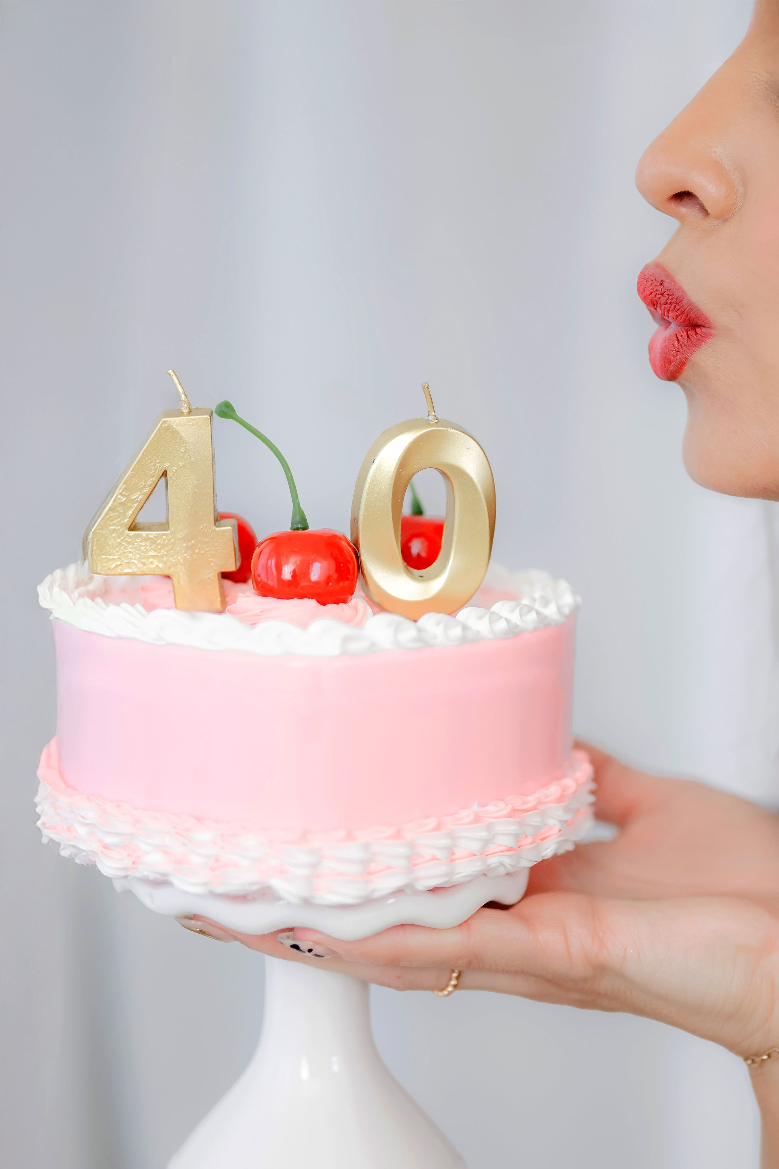 Woman blowing out gold number 40 candles on a pink birthday cake decorated with cherries on a white cake stand.