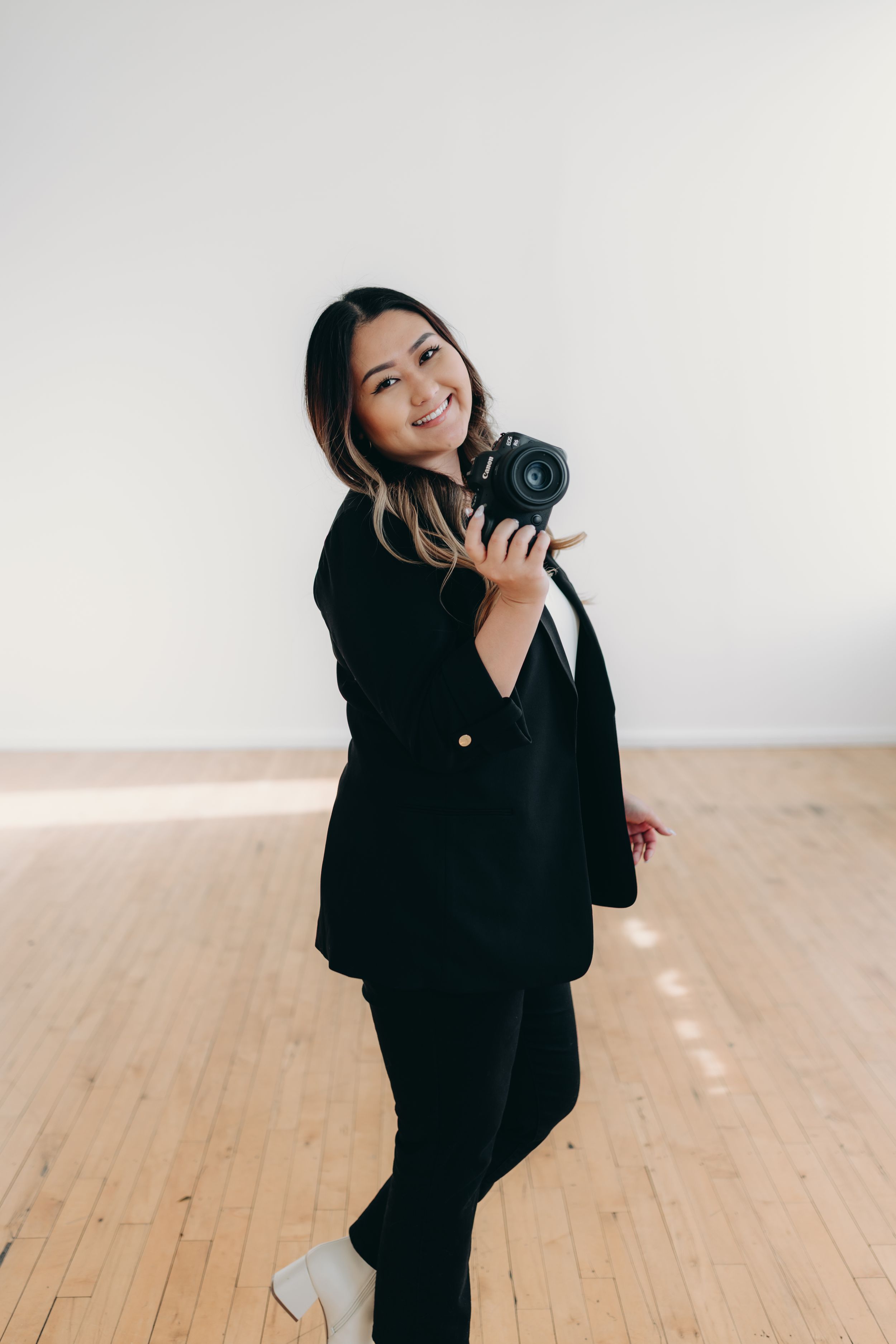 Smiling woman in black blazer holding a professional camera, standing in a bright studio with wooden floors.