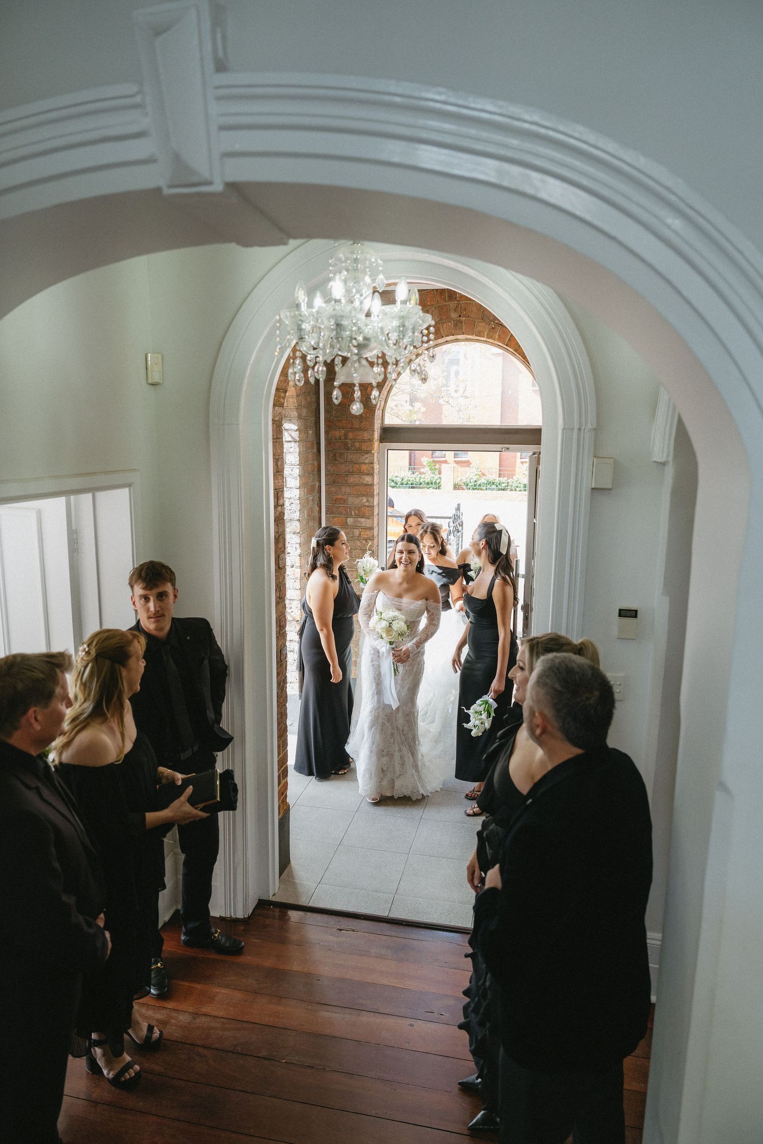 Bride in white gown walks through arched doorway flanked by bridesmaids in black dresses, guests watching in elegant venue.