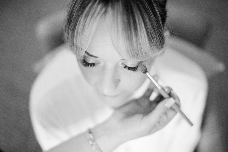 Black and white photo of a bride getting makeup applied with a brush before her wedding ceremony.