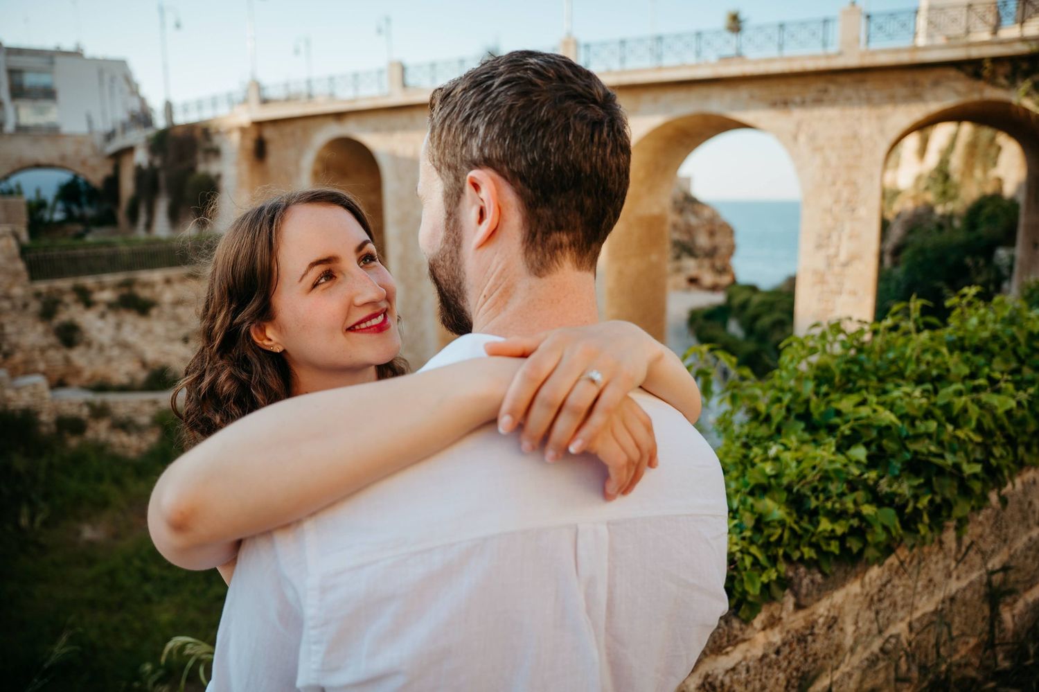 Man proposing to surprised woman in white dress by the sea with a sailboat and coastal cliffs in background.