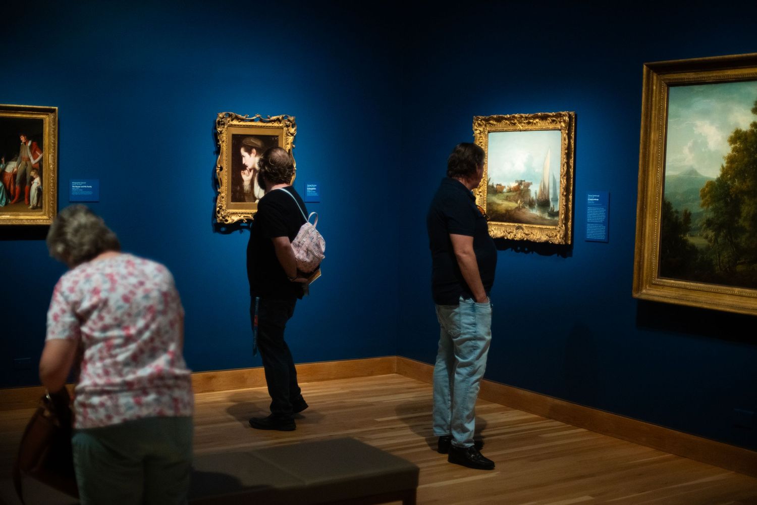 Visitors viewing classic paintings on blue walls in an art museum gallery.