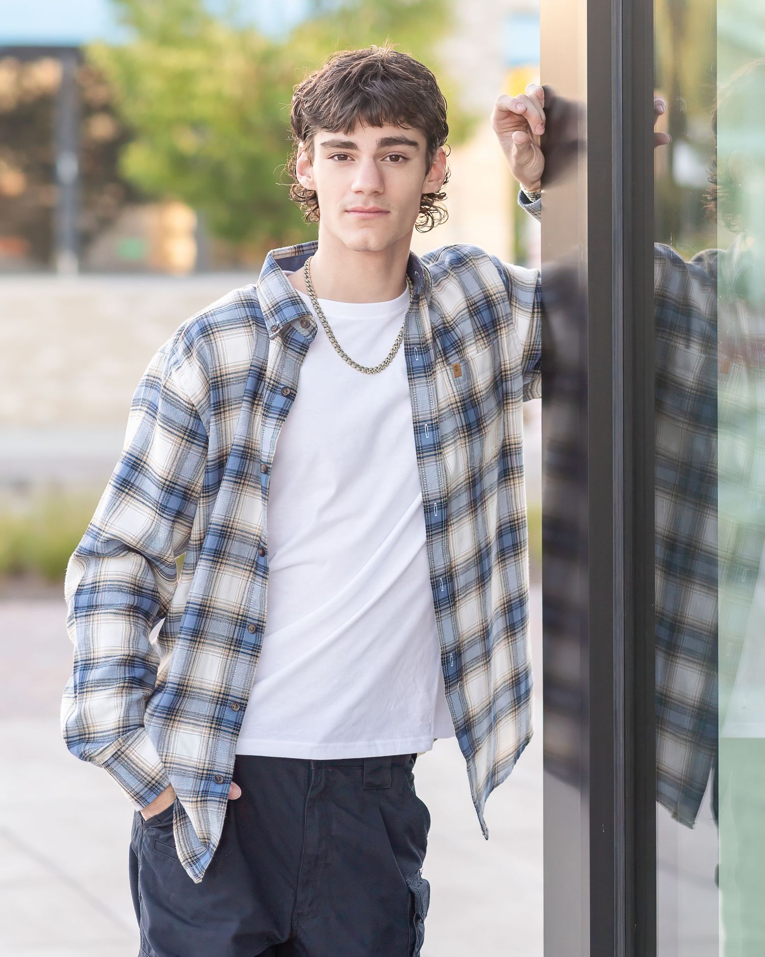 Young man in plaid shirt and Nike sneakers sitting casually against a gray shingle wall outdoors.