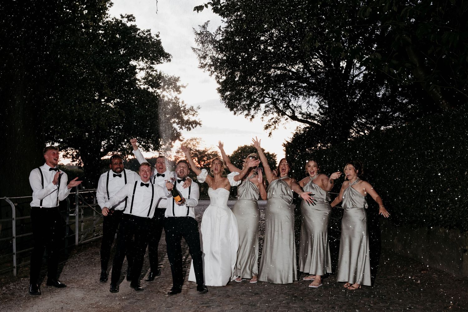 Bride and groom share a romantic dip kiss on a cobblestone path at sunset, surrounded by lush green trees.