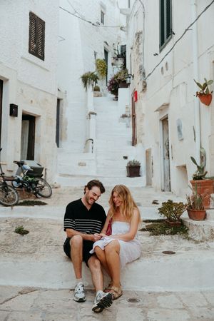 Couple sitting together on white stone steps in a charming Italian alleyway with whitewashed buildings and potted plants.