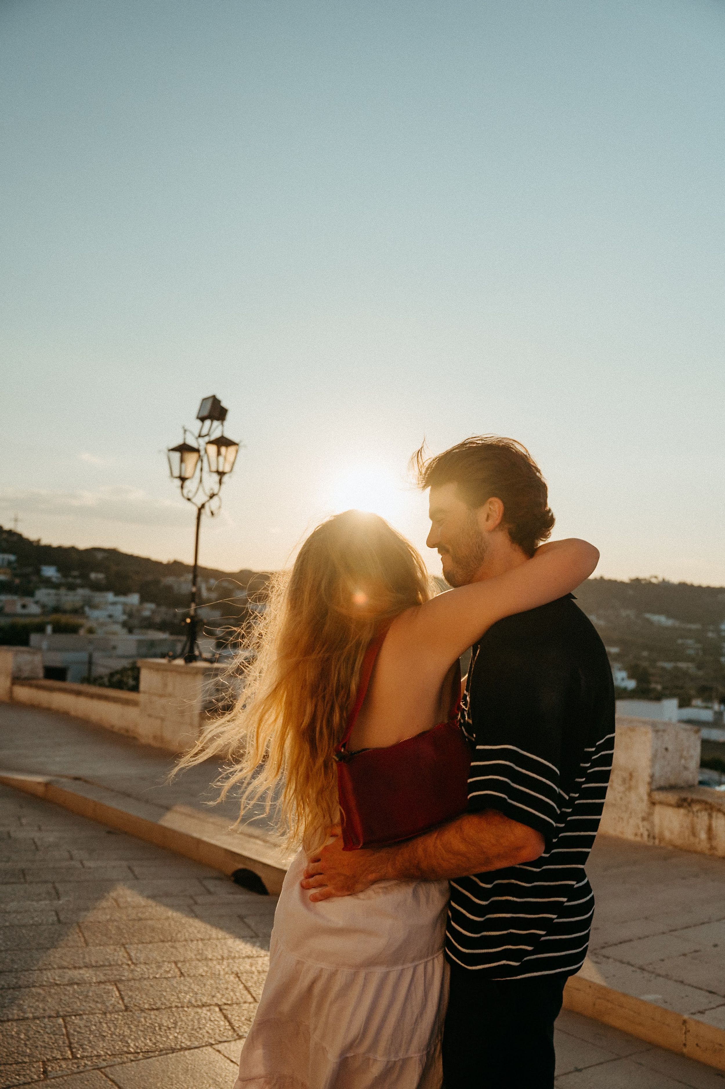 Couple embracing at sunset on a scenic terrace with a street lamp and hillside town in the background.