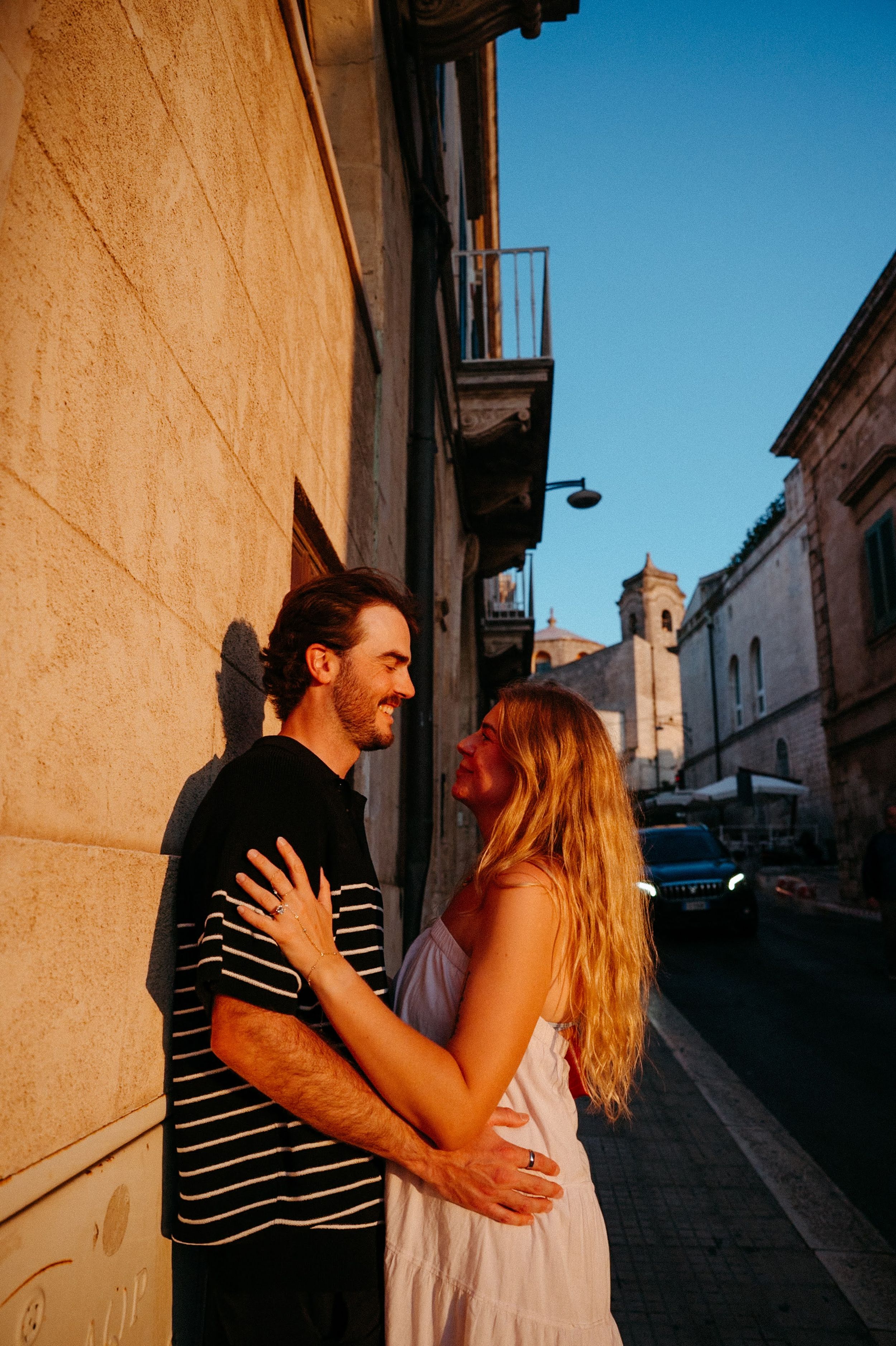 Couple embracing and smiling at each other against a sunlit wall on a charming European street at golden hour.