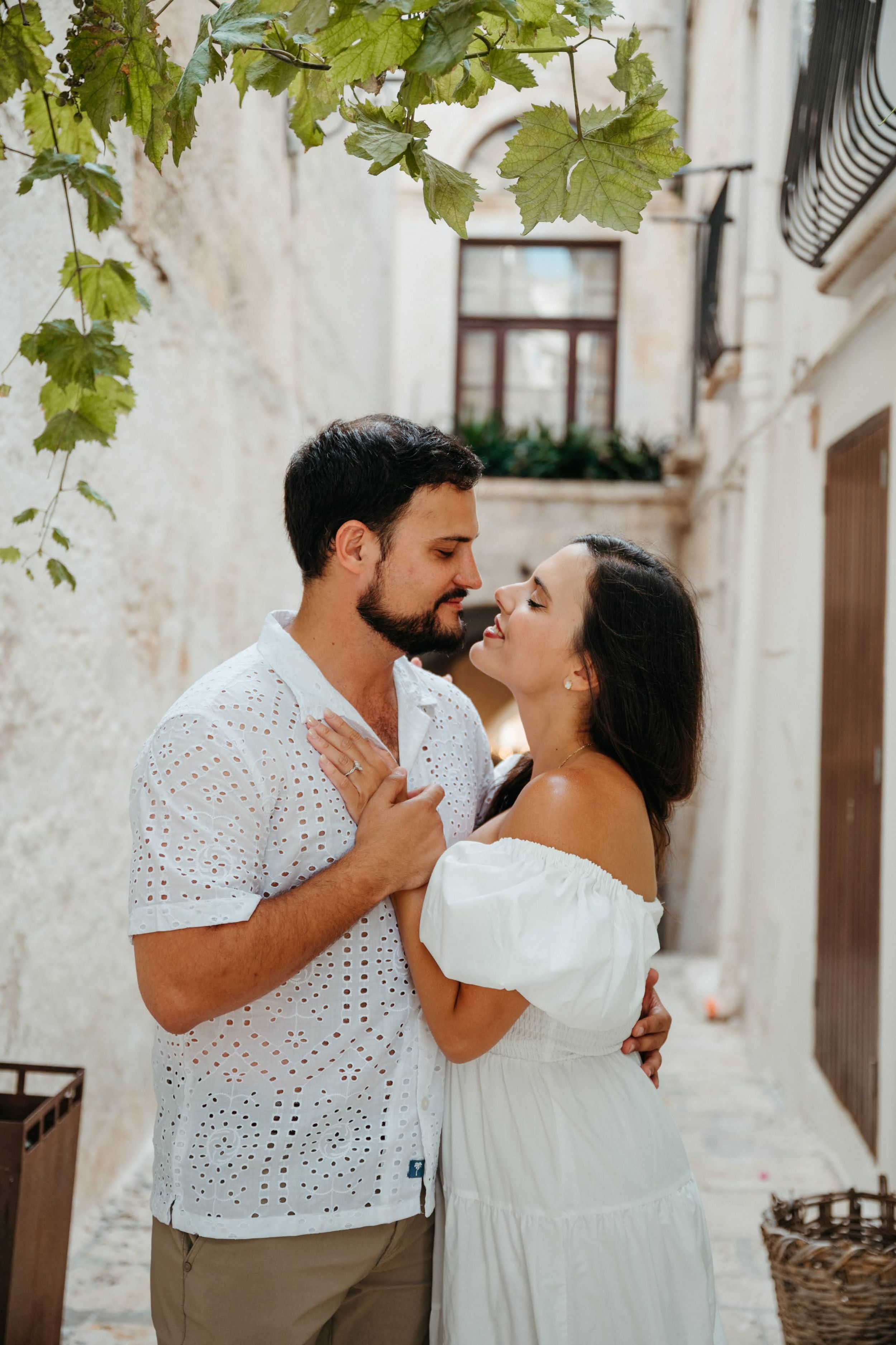 Romantic couple about to kiss in a rustic white-walled Mediterranean alley with lush green vines overhead.