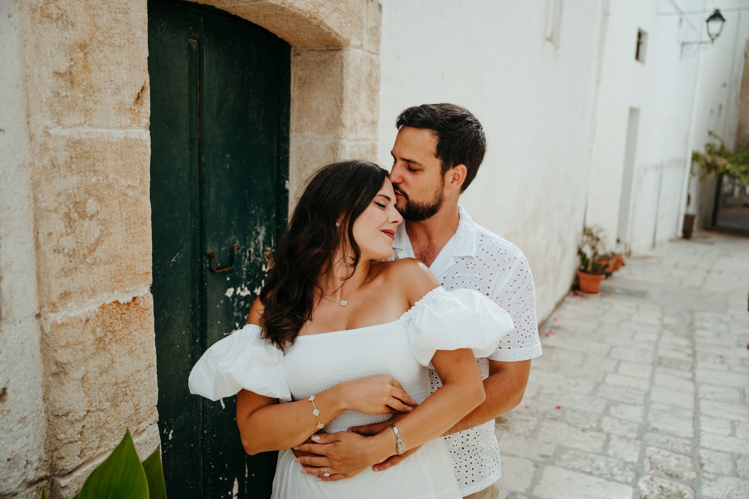 Romantic couple embracing in a charming white Mediterranean alley, woman in white dress, man kissing her cheek tenderly.