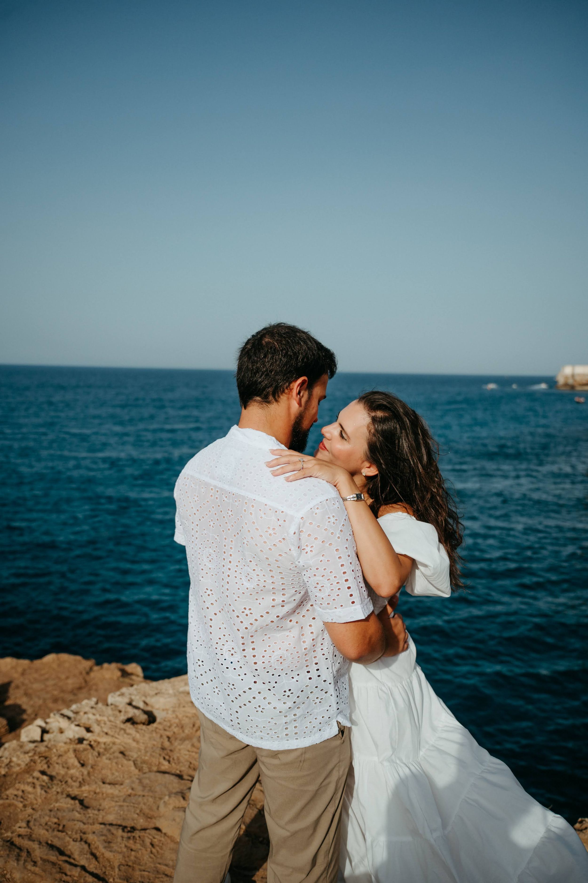 Romantic couple embracing on rocky coastal cliffs with turquoise ocean backdrop, both dressed in white outfits.