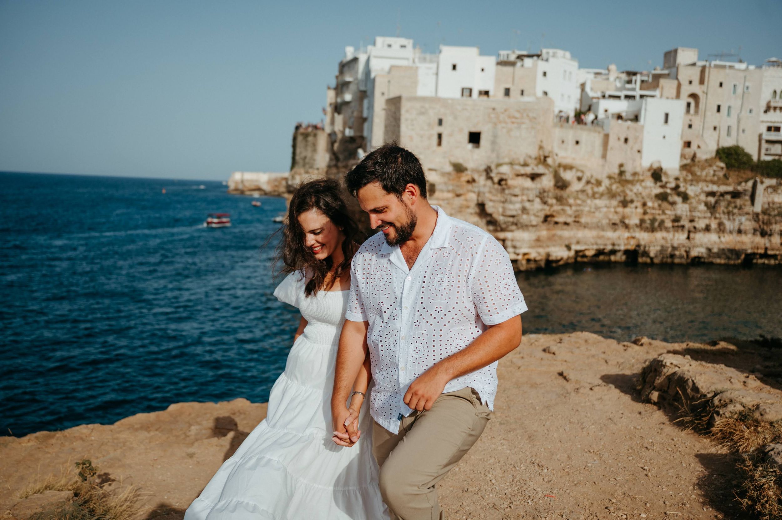 Couple holding hands on rocky cliffside with white Mediterranean coastal town and blue sea in background.