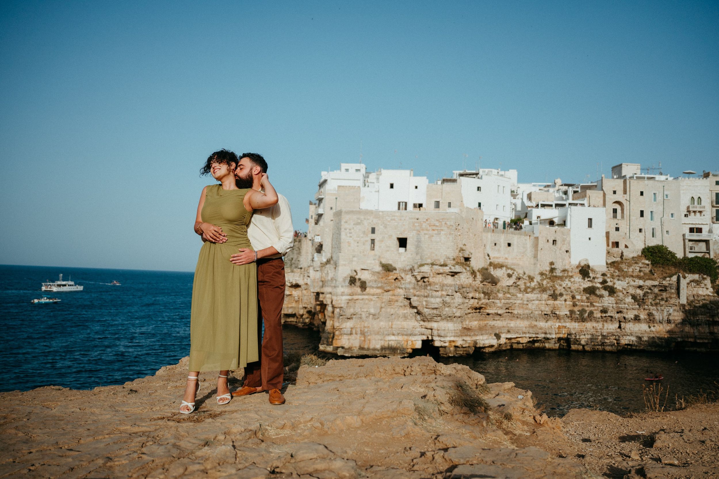 Couple embracing on rocky cliffs overlooking the white-washed coastal town of Polignano a Mare, Italy.